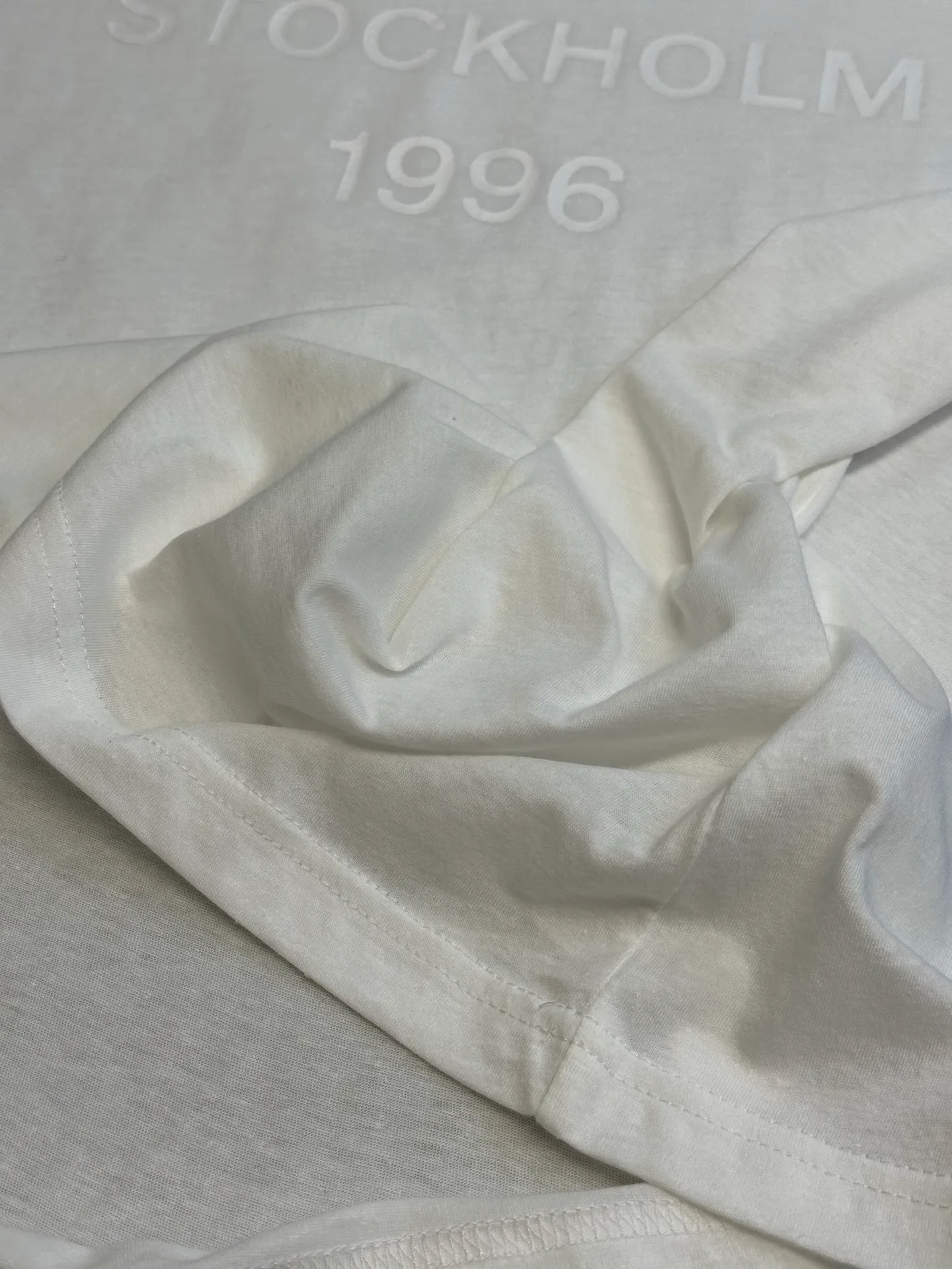 Ac White Letter Round Neck Short Sleeve The fabric is made of 32-count cotton and linen blended plain knitting, with a ratio of 85% cotton and 15% linen fiber. The weight is only 145g/m²; the ribbed neckline is made of Same Style cotton and linen blended yarn, 145g/m², to ensure a unified texture of the entire piece. The collision of the natural rough base and the industrial spraying process creates a natural irregular texture and an extremely light and breathable wearing experience. Cotton provides a skin-friendly and soft foundation, while linen introduces natural roughness and knotted particles, giving the fabric surface an irregular texture. The addition of hemp fiber makes the fabric have excellent moisture absorption and perspiration properties, while also bringing a natural micro-wrinkle texture and dry touch. After the fabric is piece-dyed, the dye evenly penetrates into the fiber, showing full color fastness and soft matte color, providing a uniform base for subsequent inkjet printing. The front is decorated with logo printing. The printing uses a double-layer printing process of environmentally friendly elastic glue and varnish, which takes into account the concept of environmental protection, wearing comfort and visual expression. The bottom layer uses environmentally friendly elastic glue, with water-based environmentally friendly resin as the base material. The color paste is scraped onto the surface of the fabric through a high-mesh screen. After drying and solidifying at high temperature, a soft and highly elastic continuous adhesive film is formed. The printed film can stretch and retract with the fabric. Even if it is covered with a large area of ​​pattern, it is still breathable and does not feel tight when wearing it. The surface layer is accurately overprinted with transparent varnish, creating a high-gloss effect in specific areas. This layer of varnish not only significantly improves the color contrast of the pattern, but also creates a local wet and bright visual and tactile contrast, making ordinary printing instantly gain three-dimensional layers and glossy texture. At the same time, the smooth surface layer also enhances Wear-Resistant and wash-resistant properties. This T-shirt is defined as a neutral style, casual fit, and hip length; its style logic pursues a genderless sense of wrapping and extended proportions. The key point is that the dress is hip-length, not only to cover the buttocks, but also to create a lazy, flowing silhouette by utilizing the lengthened body and the drape of the fabric. In terms of process execution, the side seams need to maintain a straight hem to prevent the long body from looking like a straight bucket. Taken as a whole, it's a hybrid that combines the functional details of workwear with the relaxed feel of high fashion. size：xs s m l